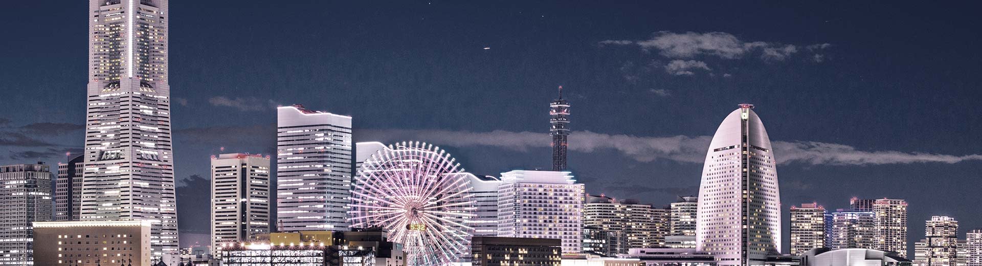 Una fila de rascacielos modernos por la noche en Yokohama se ilumina la oscuridad, con una rueda de la fortuna irreverente en primer plano.