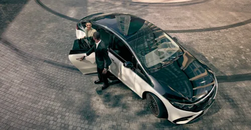 An elegant woman gets out of a two-tone Mercedes Blacklane car while a chauffeur holds the door.