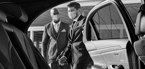 Real Blacklane chauffeur Douglas opens the door to the backseat of his vehicle for a guest in a suit, and they are both wearing face masks.