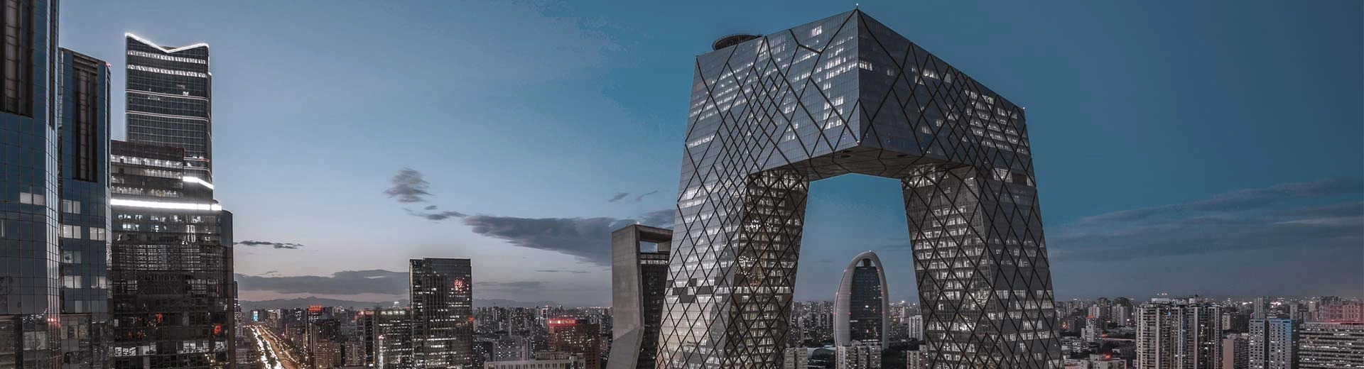 Beijing skyline at sunset, with the CCTV headquarters in view to the right. City lights glowing and buildings to the left.