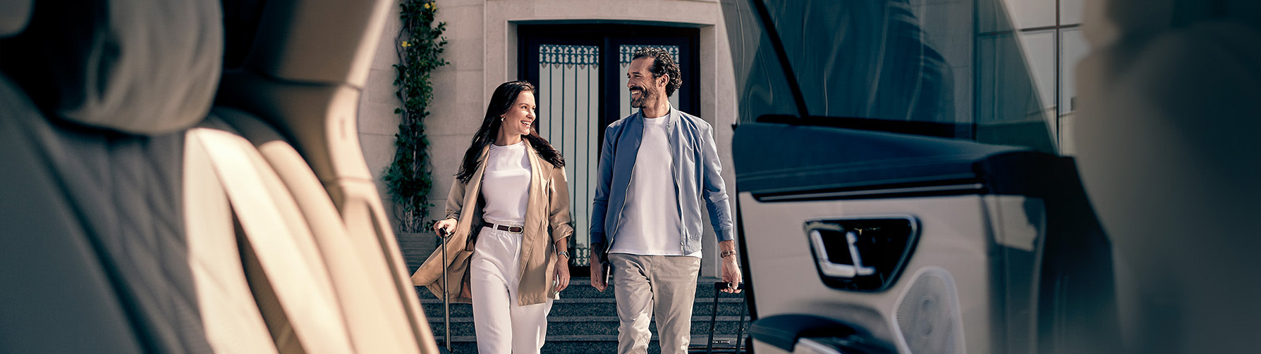 A couple with rolling luggage smile as they walk toward a Mercedes-Benz EQS.