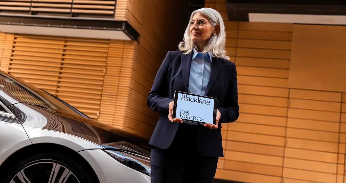 A female Blacklane chauffeur stands beside a Mercedes EQS holding a tablet with the Blacklane logo and the name "Jens Wohltorf" on it.