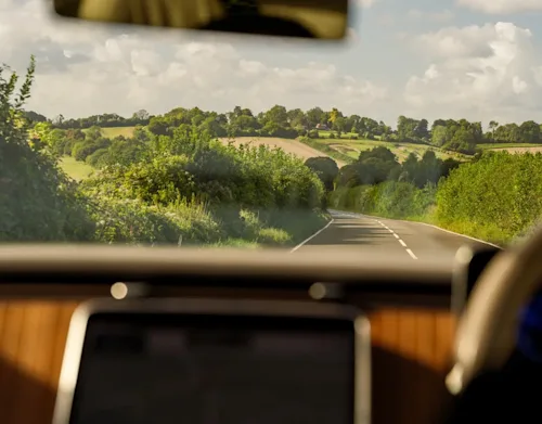 Shot of a rolling countryside road through windshield of a luxury car, showing hedges and fields.