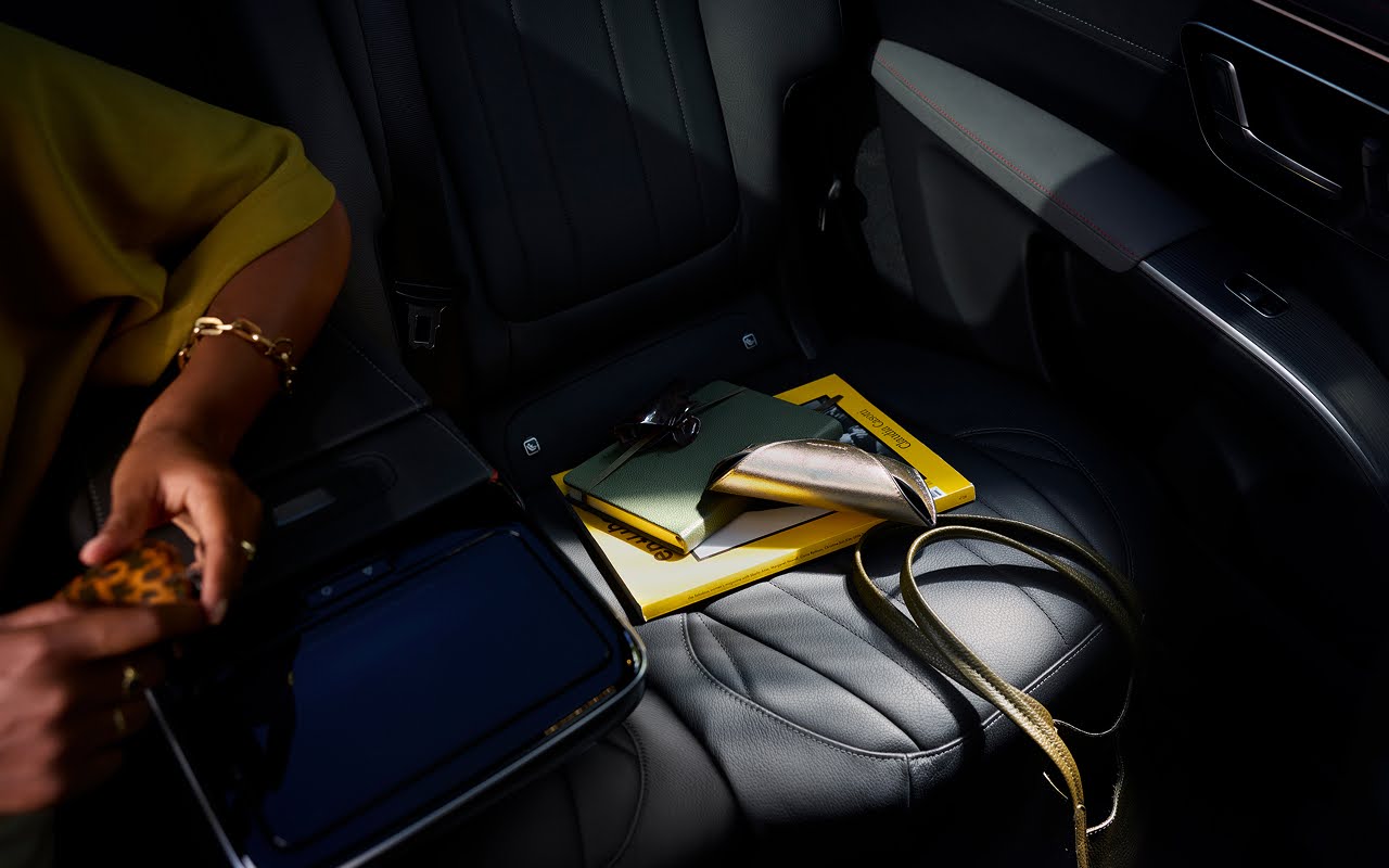 A passenger's hands in the back seat of a Blacklane limousine, with a bag, book, and sunglasses case on the passenger seat.