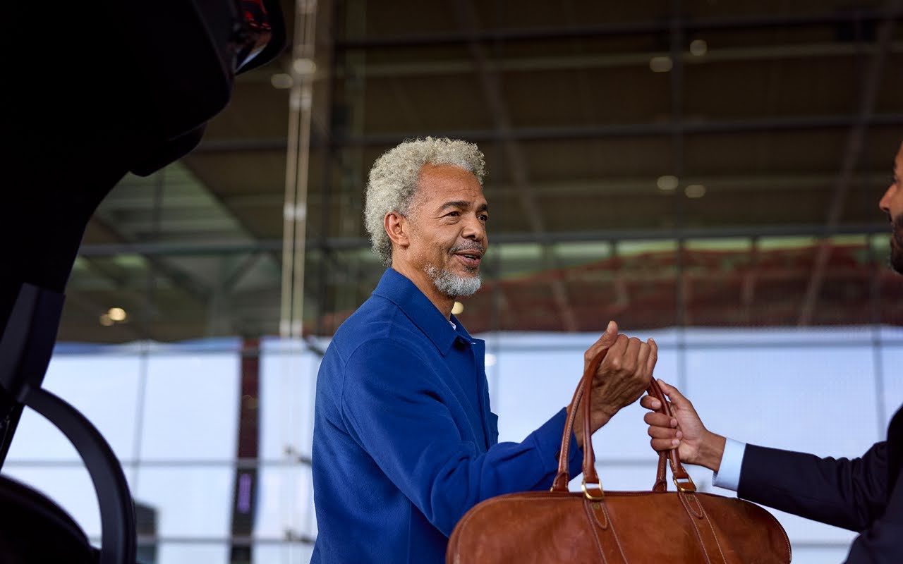 A man takes his leather bag from a chauffeur outside an airport.
