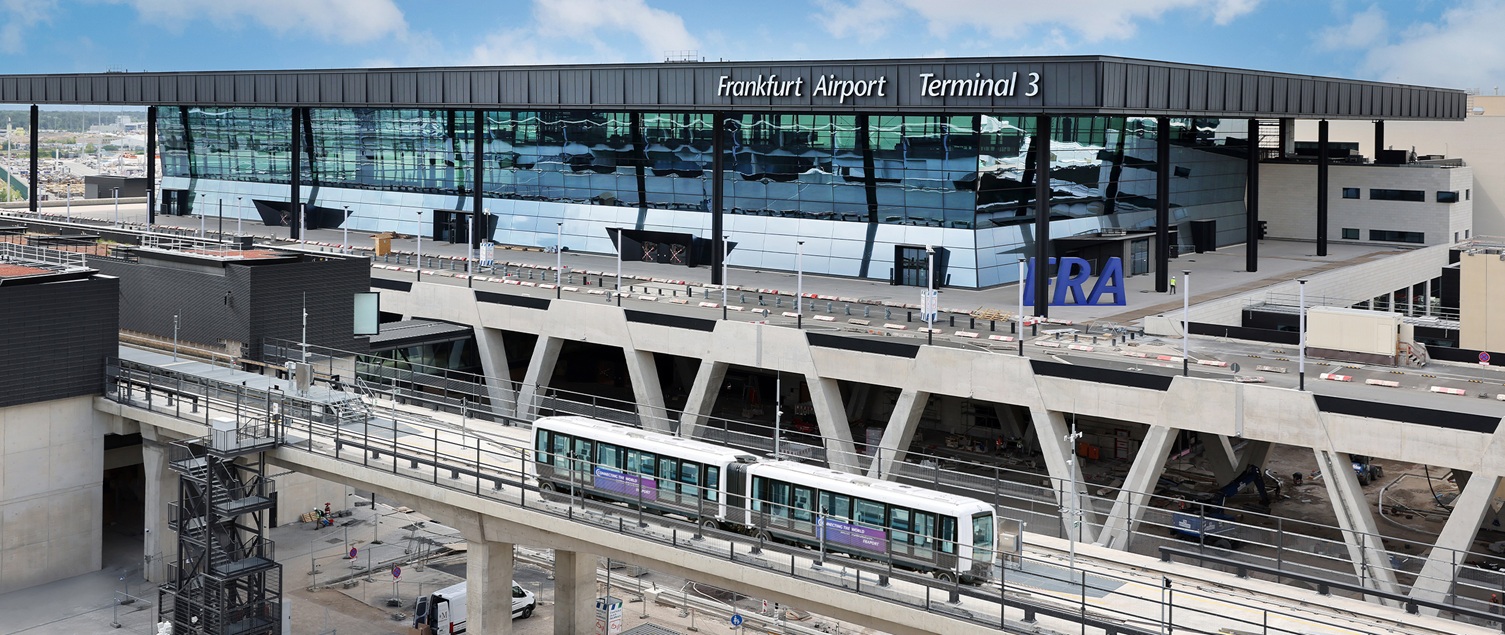 Exterior shot of the new Terminal 3 building at Frankfurt Airport, with a passenger shuttle passing by on a track in front of the building.