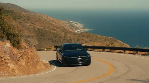 A black Mercedes EQE drives up a hill winding along the coast, with the ocean and a seaside town in the background.
