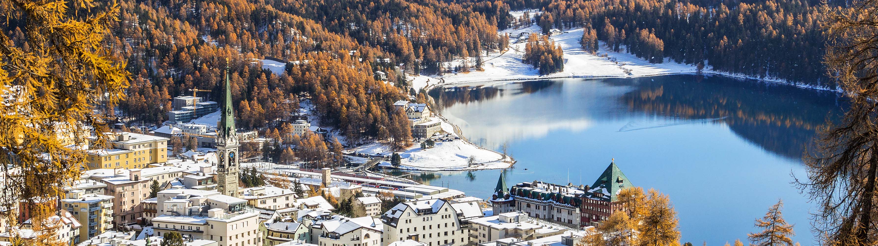 The beautiful, snow-covered village of St. Moritz sits beside a body of water on a clear day.