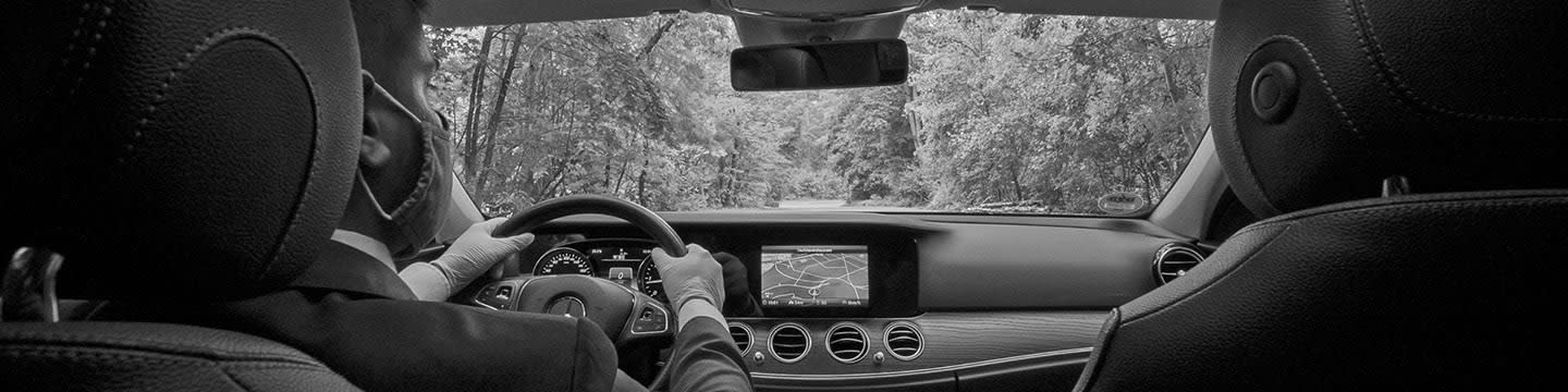 The view from the backseat of a high-class car, including a chauffeur wearing a mask, the high-tech dashboard, and the tree-lined road ahead.