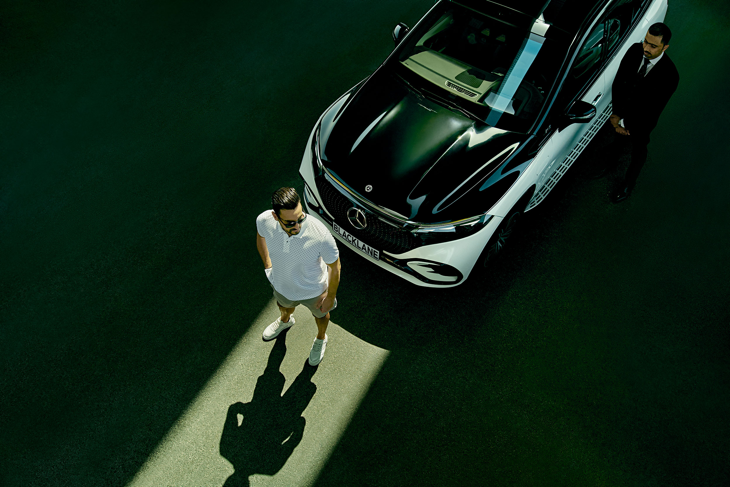 Overhead view of a guest standing in a sliver of sunlight in front of a two-toned Mercedes EQS with the chauffeur standing off to the side.