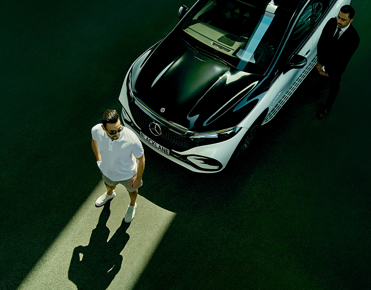 Overhead view of a guest standing in a sliver of sunlight in front of a two-toned Mercedes EQS with the chauffeur standing off to the side.