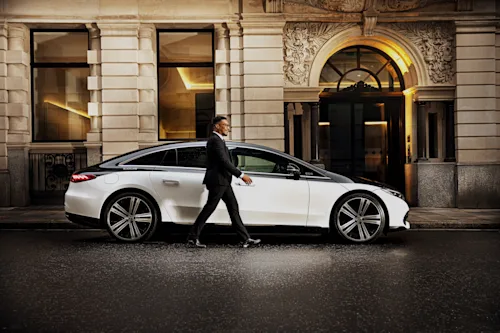 A Blacklane chauffeur in a smart suit walks beside a two-tone Mercedes EQS outside a fancy London hotel.