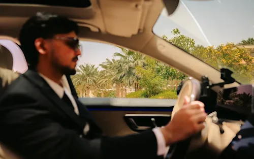 A chauffeur in a suit with sunglasses driving a luxury car, with the shot focussed on the palm tree vegetation out the driver's window.