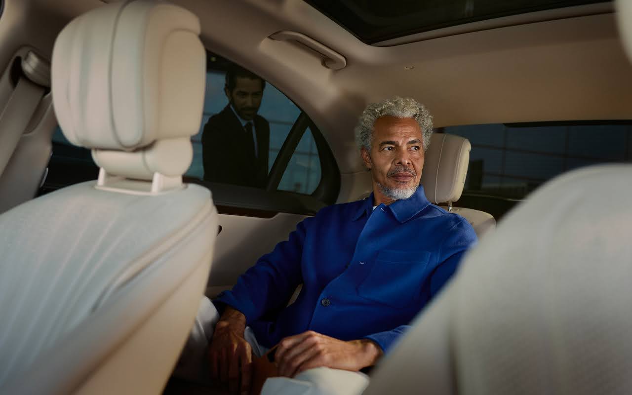 A man sits in the back seat of a Blacklane limo as a chauffeur waits to open the door outside.