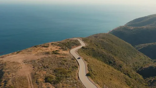 Overhead of a black SUV driving over a windy coast road with the ocean in the background.