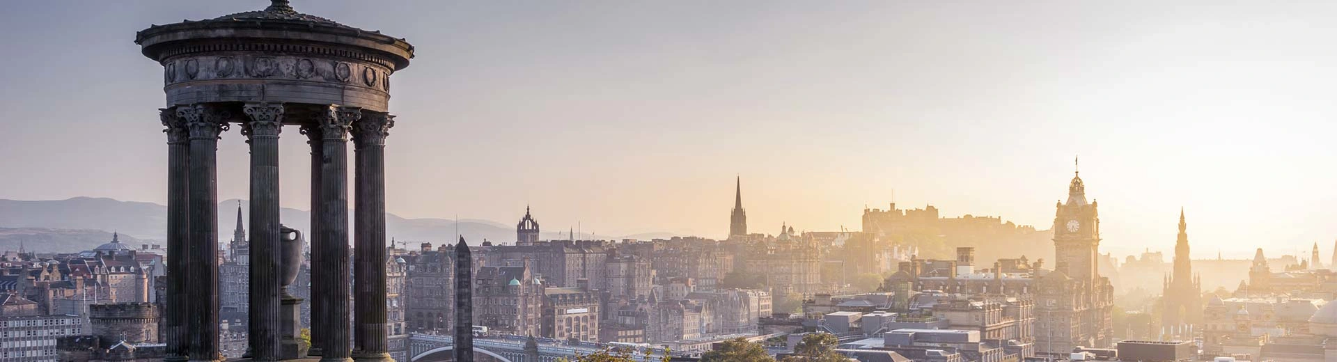 The sun pierces the sky over the skyline of Edinburgh, with the castle silhouetted in the background.