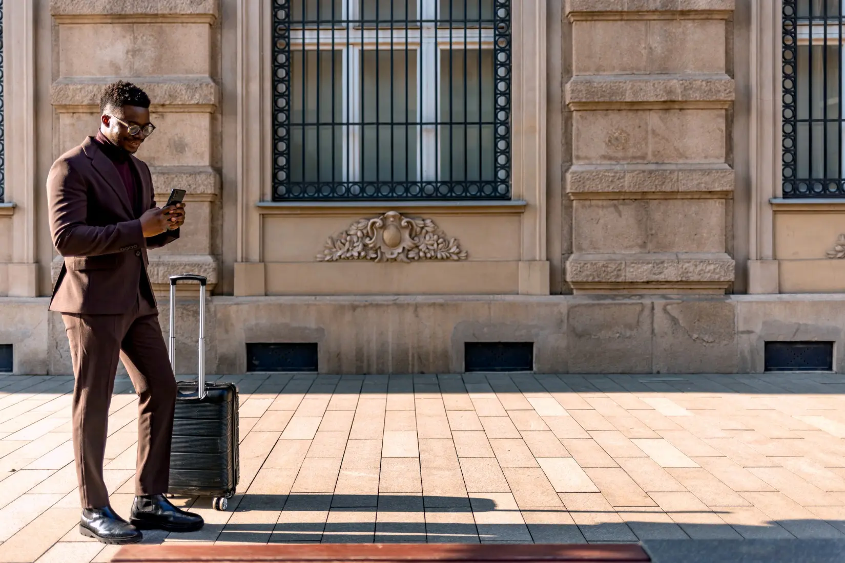 A businessman in a suit with a suitcase looking at his phone standing in front of a building after landing at his business travel destination.