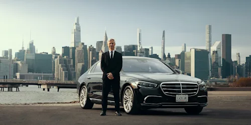 A Blacklane chauffeur stands in front of his black Mercedes limo with the East River and the skyscrapers of Manhattan in the background.