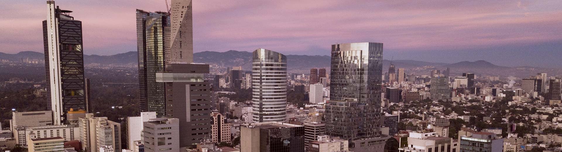 前景に高層ビル群、後景にメキシコシティが広がり、背景には夕暮れや夜明けの紫色の光。