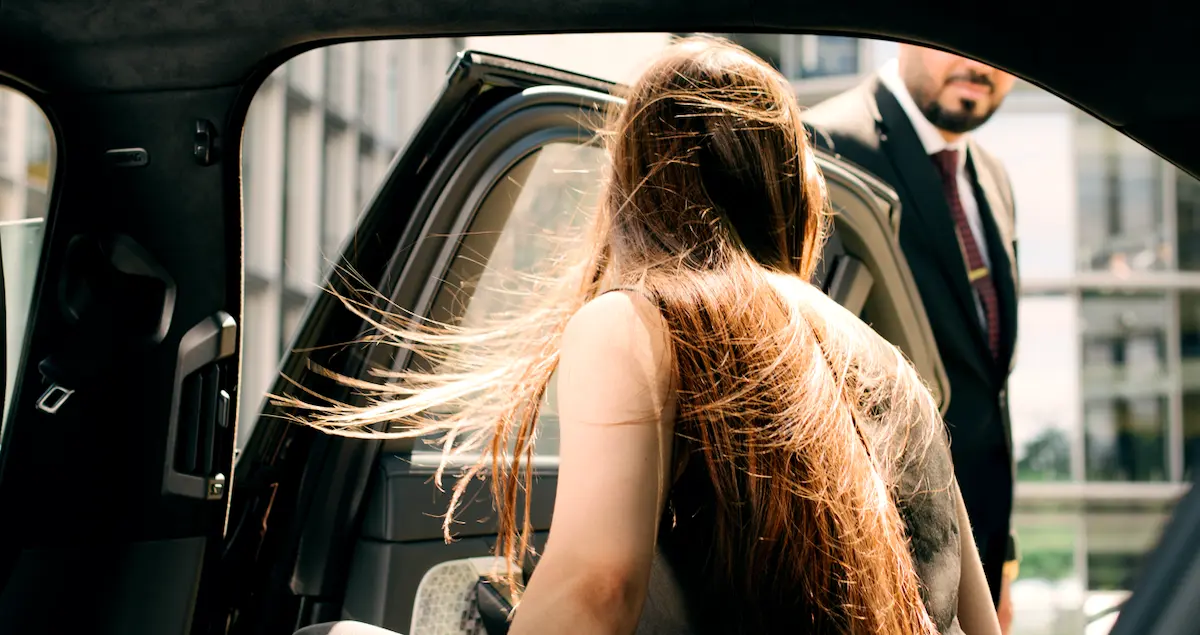 Shot of the back of a glamourous woman getting out of a Blacklane as a chauffeur holds the door open, her hair blowing in the wind.