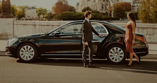 A Parisian Blacklane chauffeur holds open the door of a Mercedes limo for a woman in a dress.