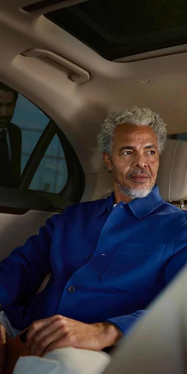 A well-dressed man with grey hair relaxes in the back seat of a Blacklane limo while a chauffeur waits outside.