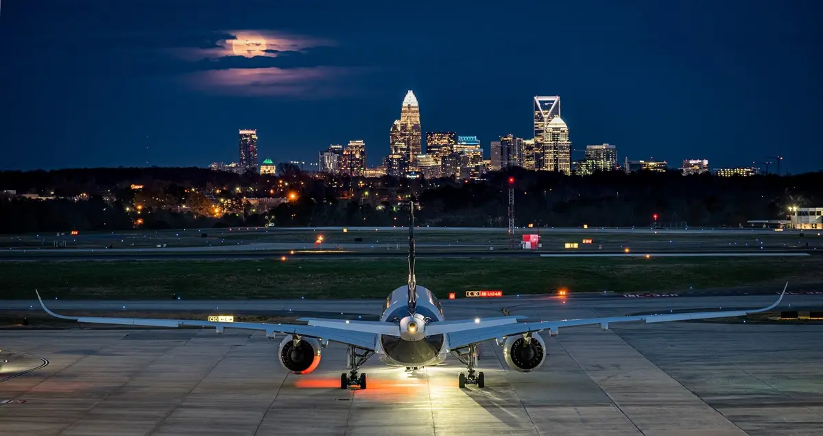 A plane on the runway at CLT Airport at night, with downtown Charlotte's buildings lit up in the background beside the moon.