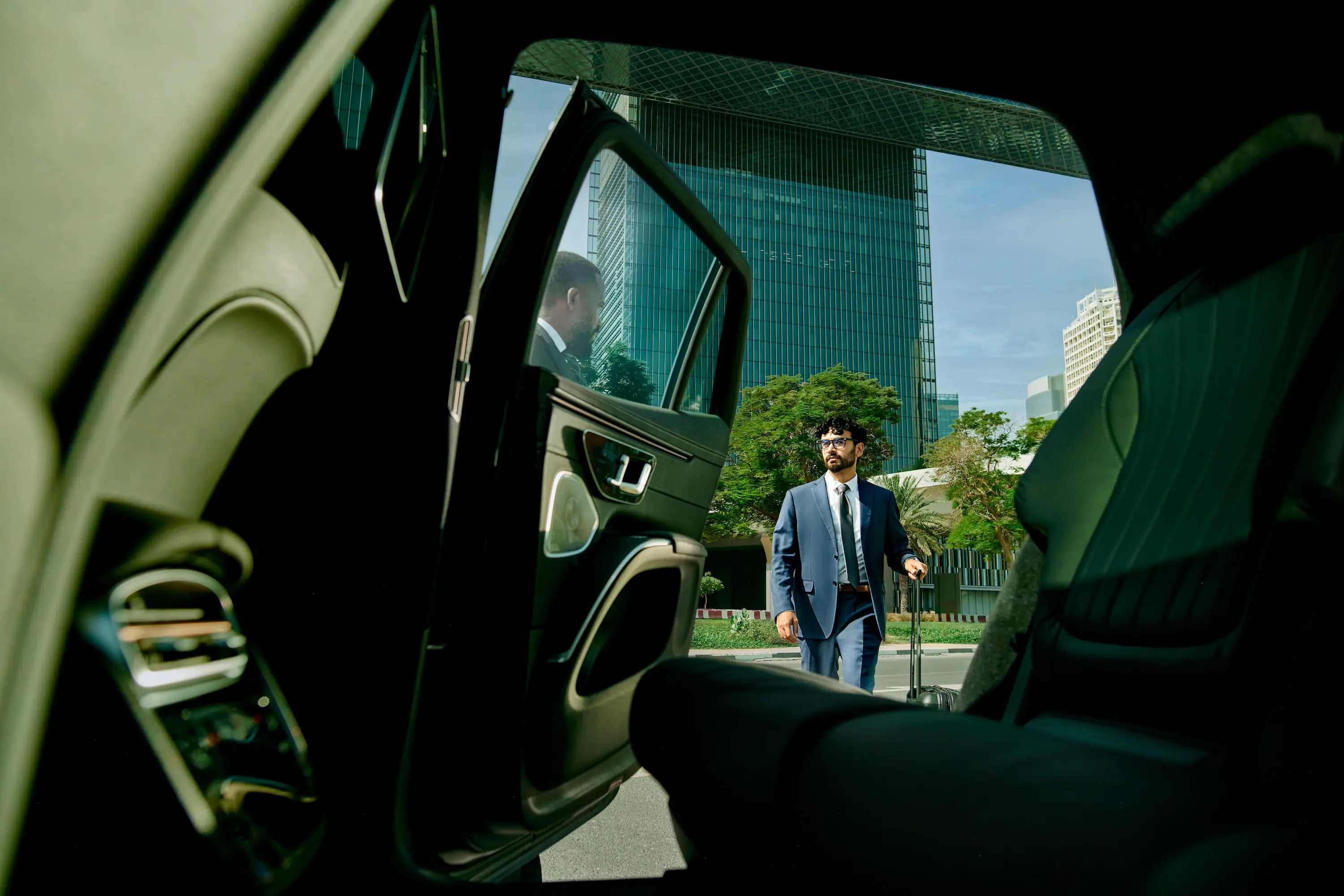 From the backseat interior of a car, a man in a suit can be seen from the open door walking towards the car with rolling luggage.