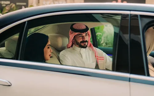From the backseat interior of a car, a man in a suit can be seen from the open door walking towards the car with rolling luggage.