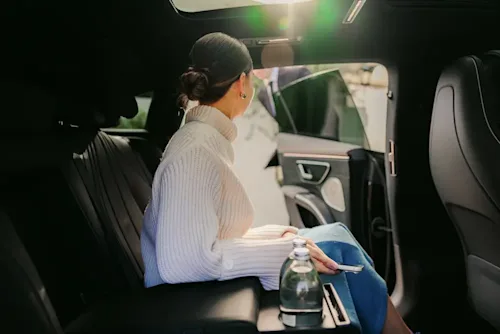 A female passenger in the backseat of a Blacklane holds her phone while looking out the open car door the chauffeur is holding for her.