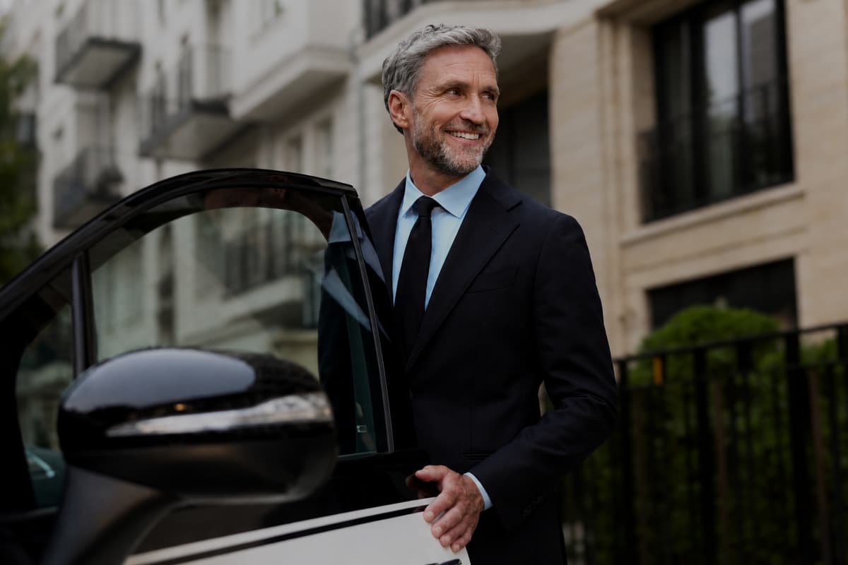 A male chauffeur smiling and about the close the door of a Mrecedes-Benz EQS.