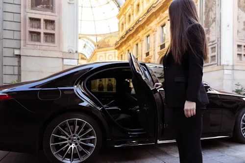 A female chauffeur holds open a black limo door with an ornate Milanese building in the background.