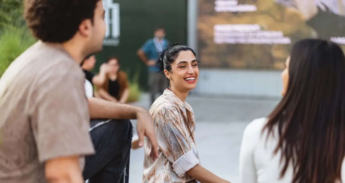 A young woman in a Dubai members club laughs as she sits in a courtyard with her acquaintances, a man and a woman.