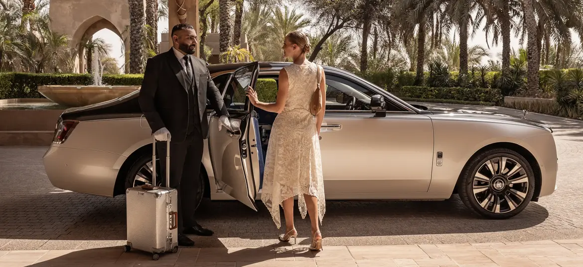An elegant woman is greeted by a Blacklane chauffeur beside a two-tone Rolls Royce Phantom outside a hotel in Dubai.