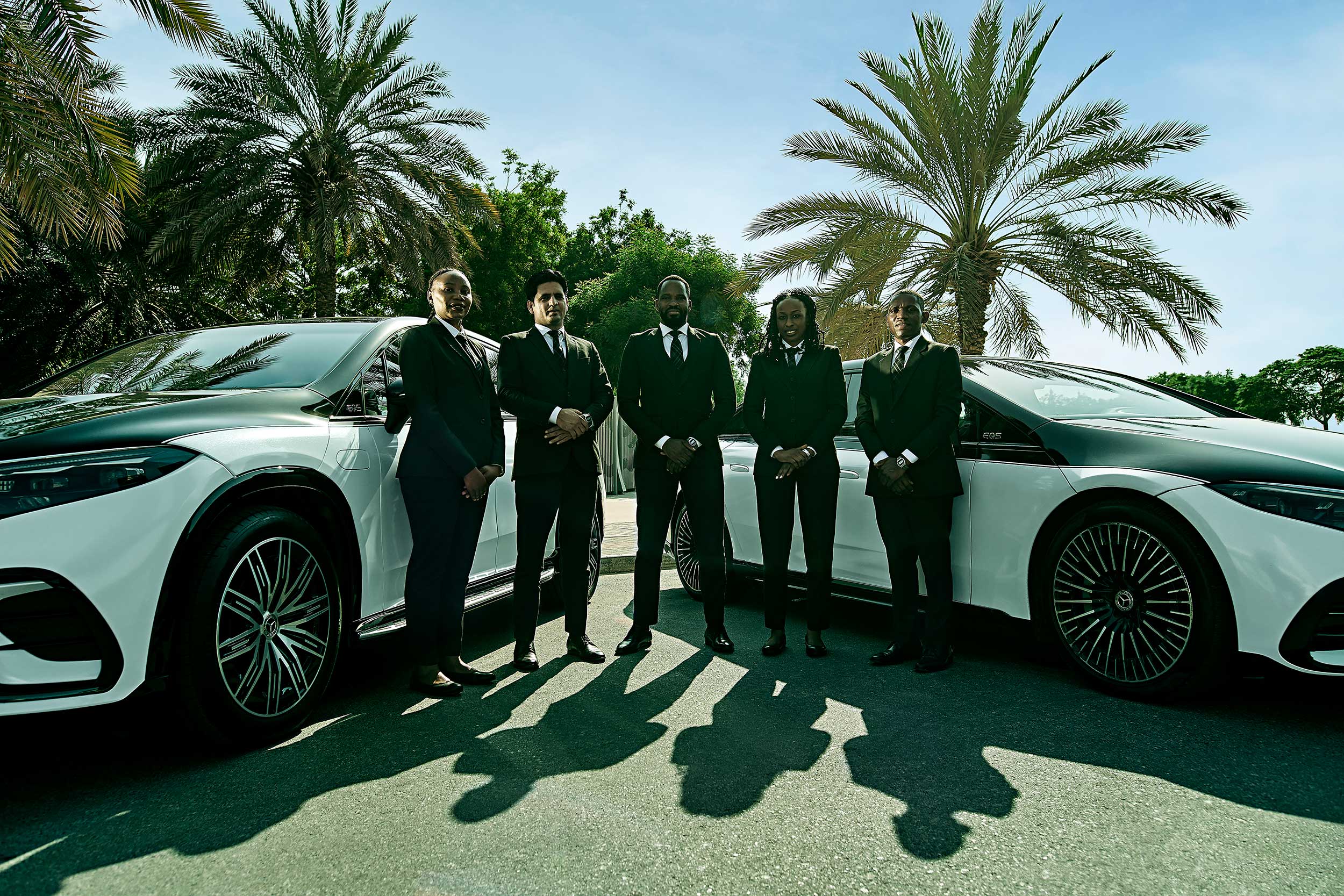 Five chauffeurs standing in a line in front of two, two-toned Mercedes EQSes with palm trees in the background.