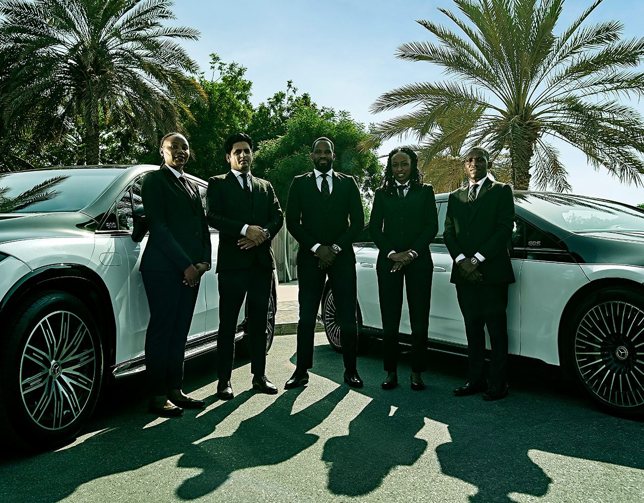 Five chauffeurs standing in a line in front of two, two-toned Mercedes EQSes with palm trees in the background.