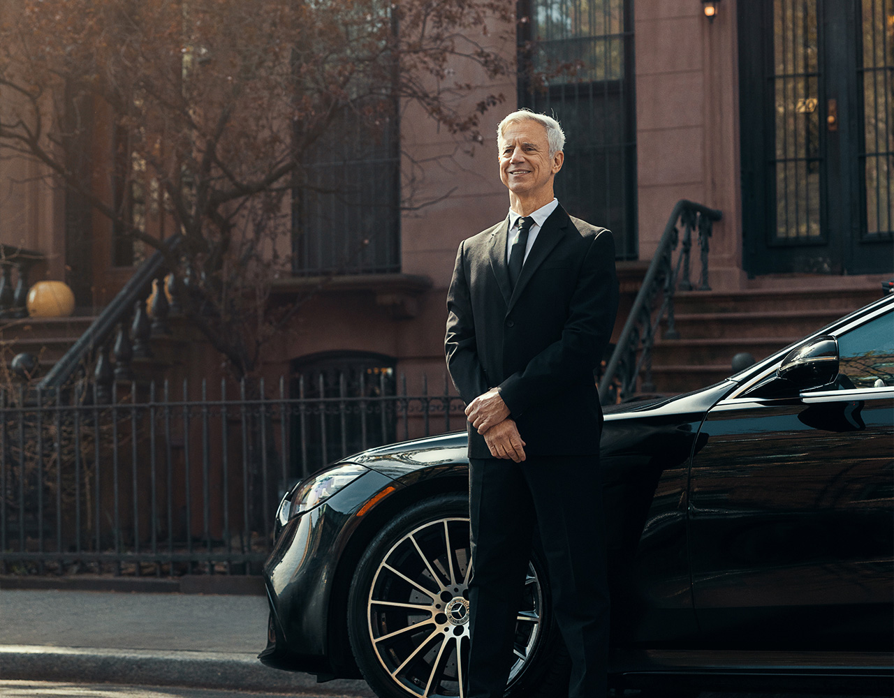 A smiling and cleancut chauffeur stands beside his stylish vehicle.