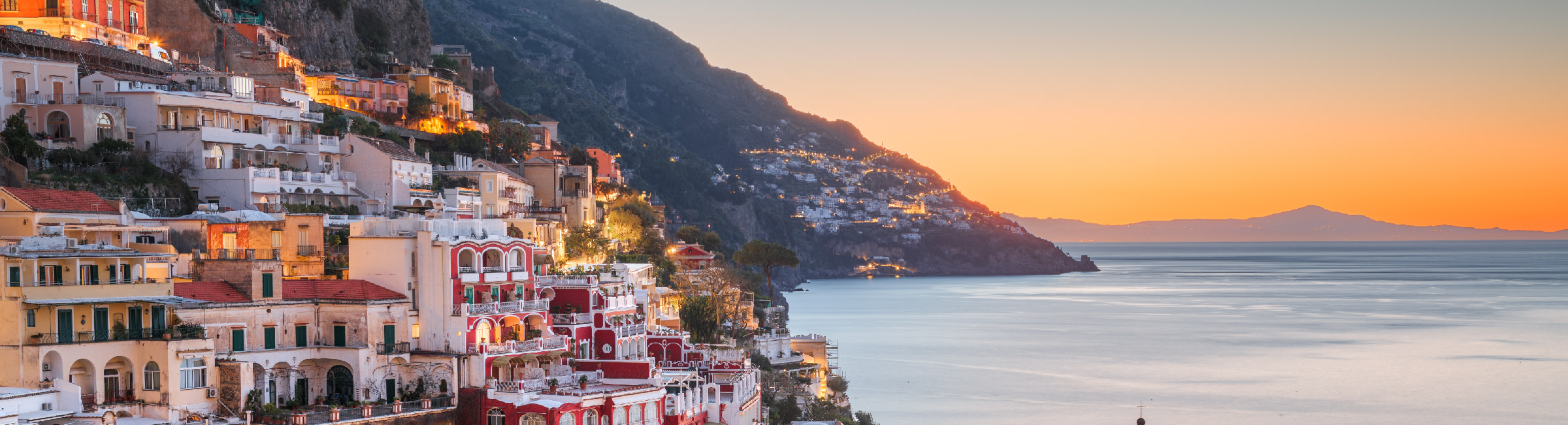 Small white houses sit on hillsides overlooking the bay of Positano and a beautiful sunset.