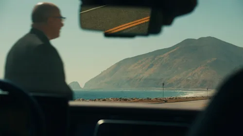 A Blacklane chauffeur in a suit walks across the front of the car, shot through the windscreen from the front seat.