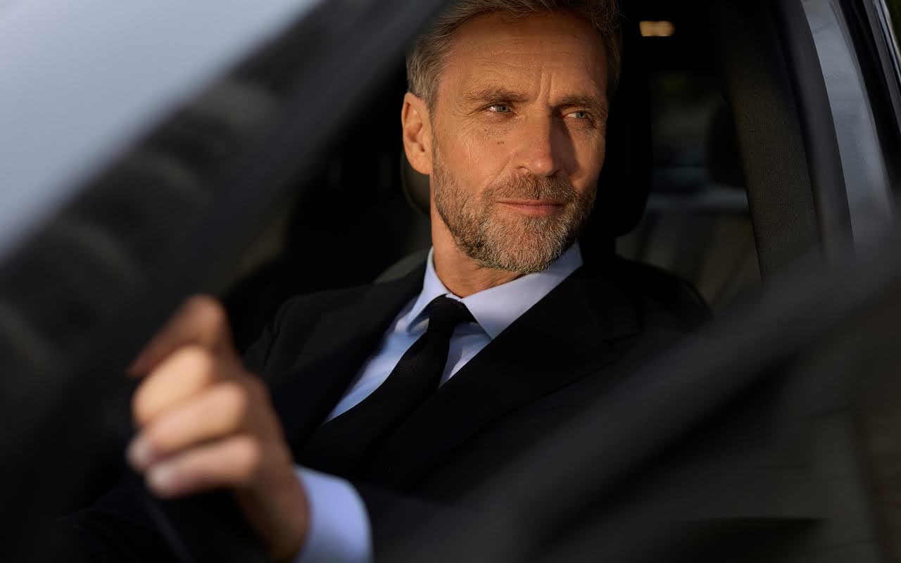 A suited chauffeur looks out the open window of the limo he is driving.