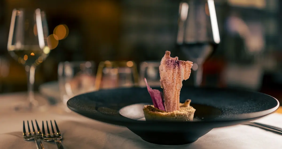 An elegantly displayed plate of fine food with a tuile sticking straight out of it on a private dining table in a top restaurant, with red wine glasses in the background.