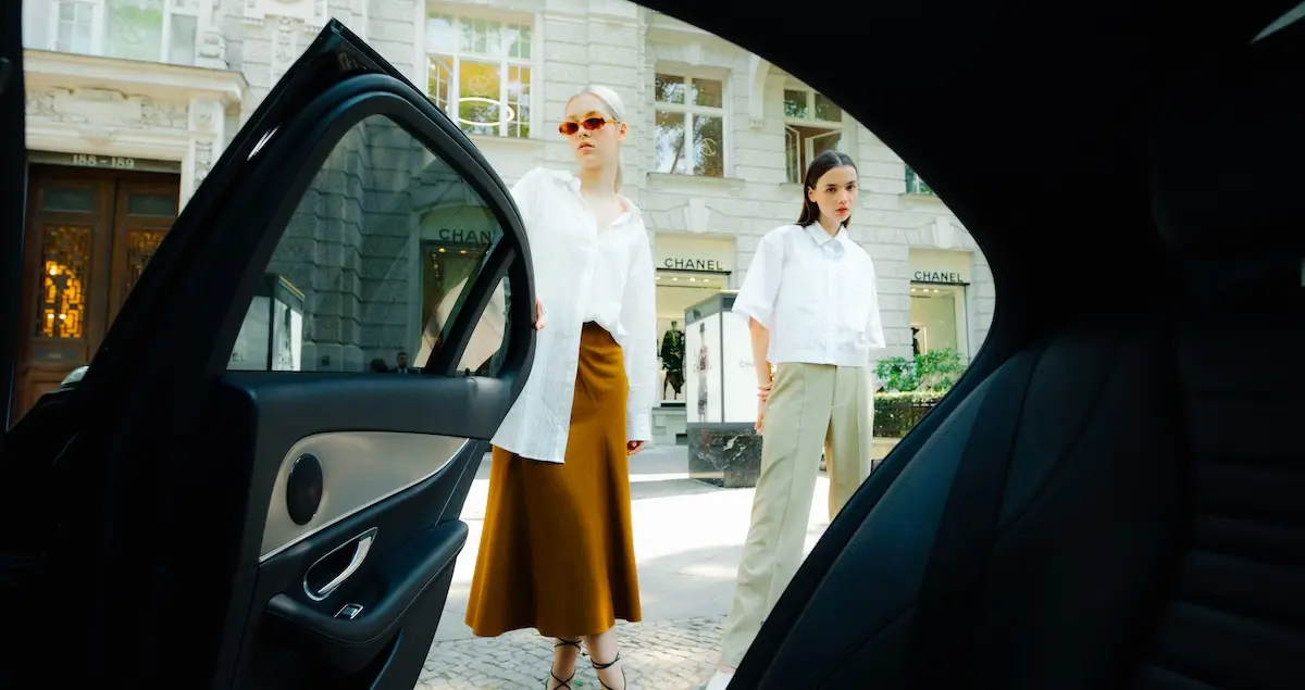 Two fashion models pose standing on a European street with a Chanel store behind them, shot through the backseat door of a car.