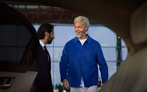 A passenger in a blue shirt gets into a Blacklane limo as a chauffeur holds the door open for him.