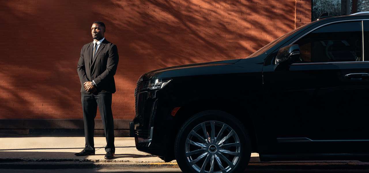 A Blacklane chauffeur in a suit stands beside a sleek car with his hands folded in front of him.