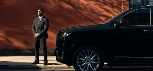 A Blacklane chauffeur in a suit stands beside a sleek car with his hands folded in front of him.