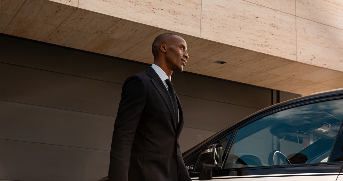 A Blacklane chauffeur in a sharp suit approaches the driver's door of his Mercedes EQS limousine.