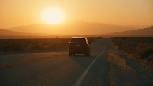 A black SUV drives away from camera down a straight desert road at sunset.