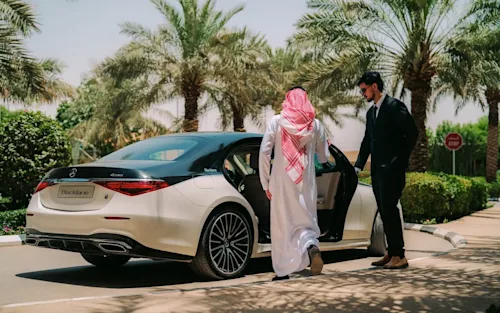 Shot from behind of a man in traditional Saudi dress entering a Mercedes EQS limo while the suited chauffeur holds the door open.