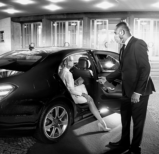 Real Blacklane chauffeur Ramanan helps a well-dressed woman get out of his vehicle parked in front of a venue. Both wear face masks.