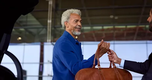 A male traveller with grey hair takes his bag from a Blacklane chauffeur outside an airport terminal building.
